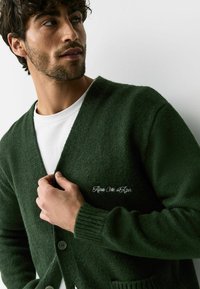 Groene gebreide cardigan met een V-hals, knoopsluiting en geribbelde manchetten. Voorzien van geborduurde tekst op de borst. Draagt over een wit T-shirt.