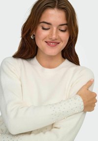 Maglione color crema con collo rotondo, realizzato in materiale morbido. Maniche decorate con perle e strass di varie dimensioni.