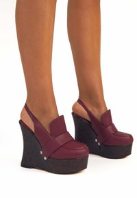 Scarpe a zeppa bordeaux con una tomaia in pelle elegante, aperte sul retro e con tacco a piattaforma. Base in legno scuro con texture e accenti argentati.