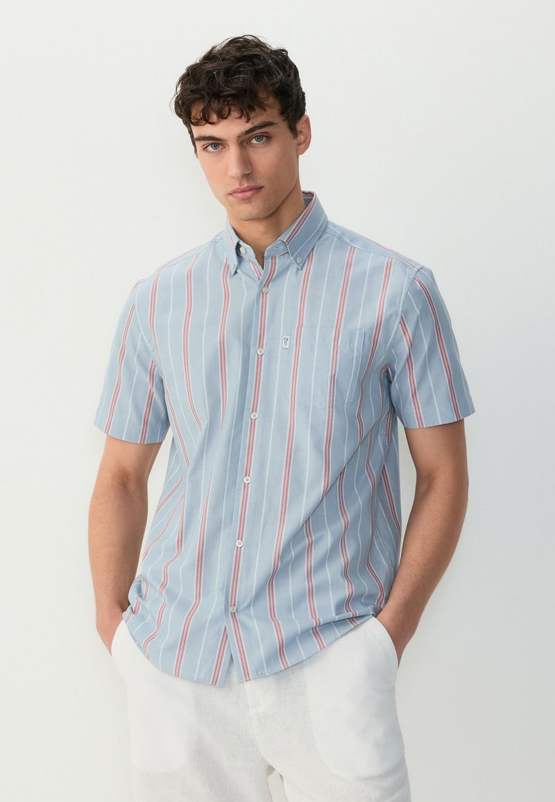 Košulja kratkih rukava na kopčanje u svijetloplavoj boji s okomitim crvenim i bijelim prugama, s lijevim džepom na prsima i ovratnikom.