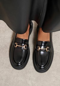 Černé lakované kožené mokasíny s tlustou černou podrážkou, zdobené zlatým řetízkem přes přední část. Hladká a lesklá textura.