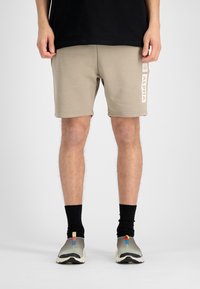 Beige sporta šorti, izgatavoti no mīkstas auduma, ar gaismas rozā "ALPHA" logotipu sānos. Valkāti ar melnām zeķēm un sporta apaviem.