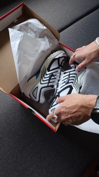 Pantofi sport albi cu accente negre, parte superioară din plasă, talpă texturată și un mic detaliu albastru, așezați într-o cutie de carton cu hârtie de ambalaj.