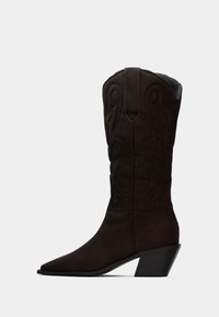 Καφέ σουέτ καουμπόικο μποτάκι με διακοσμητικά ραφές, μυτερή μύτη και κεκλιμένη τακούνι σε στοιβάδα, σε ανοιχτό γκρι φόντο.