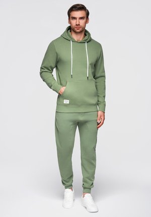 Mężczyzna stojący, ubrany w jasnozieloną bluzę z kapturem i sznurkami, pasujące spodnie dresowe oraz białe sneakersy, jedna ręka w kieszeni bluzy, neutralne tło.