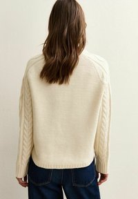 Suéter de punto color crema con un patrón de trenzas texturizado a lo largo de las mangas, dobladillo de canalé y corte relajado. Pantalones de mezclilla oscuros visibles.