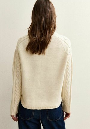 Krämfärgad stickad tröja med ett texturerat kabelmönster längs ärmarna, ribbad nederkant och avslappnad passform. Mörka jeans synliga.