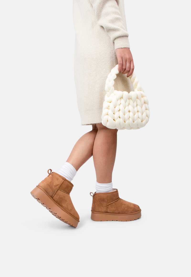 Botas de ante marrón hasta el tobillo con suela gruesa, combinadas con calcetines blancos acanalados y un bolso de crema tejido. Vestido beige liso.