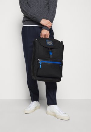 Sac à dos noir avec fermeture éclair bleue, bretelles réglables et détail de logo sur le rabat. Confectionné en tissu résistant, il présente une poche avant et un design structuré.