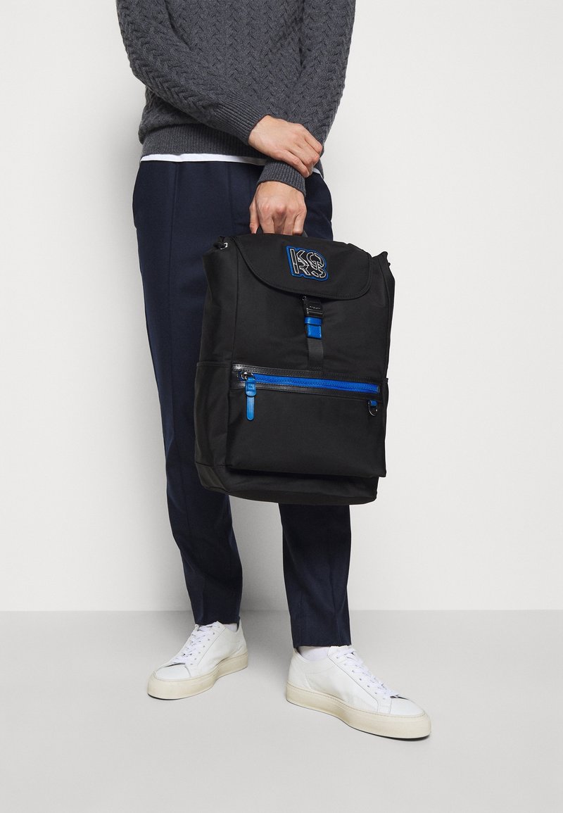 Sac à dos noir avec fermeture éclair bleue, bretelles réglables et détail de logo sur le rabat. Confectionné en tissu résistant, il présente une poche avant et un design structuré.