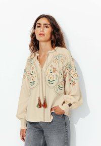 Blouse beige brodée avec des motifs floraux, une patte de boutonnage avec des franges, et de longues manches bouffantes avec des poignets élastiques, assortie à un jean gris.