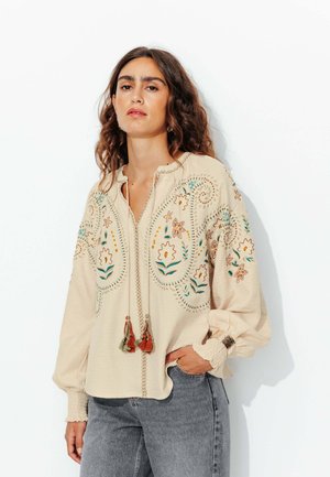 Blouse beige brodée avec des motifs floraux, une patte de boutonnage avec des franges, et de longues manches bouffantes avec des poignets élastiques, assortie à un jean gris.