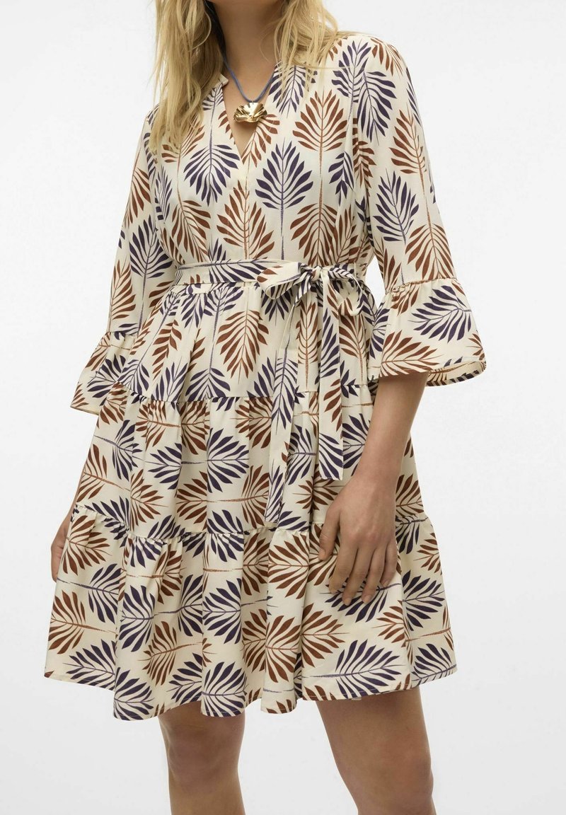 Femme portant une robe crème avec un motif de feuilles marron et violet, manches 3/4 à volants, jupe à étages et ceinture nouée à la taille.