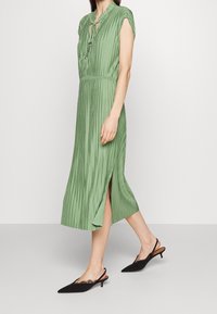 Grünes, plissiertes Midikleid mit einem Schlüssel-ausschnitt und kurzen Ärmeln. Hat seitliche Schlitze und wird mit schwarzen, spitzen Slingback-Schuhen kombiniert.