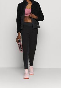 Vrouw in een zwarte fleecejack, roze sport-bh, zwarte joggers, roze sneakers die een roze Puma-waterfles vasthoudt, staande tegen een lichte muur.