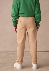 Pantalon beige à coupe ajustée, doté de deux poches arrière à boutons, associé à un sweat-shirt vert et des baskets blanches.