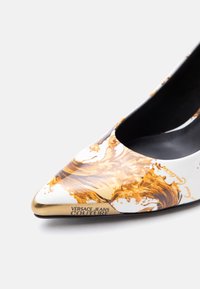 Λευκή μυτερή ψηλοτάκουνη γόβα με χρυσά μπαρόκ σχέδια και τη σήμανση "Versace Jeans Couture" στη μύτη.