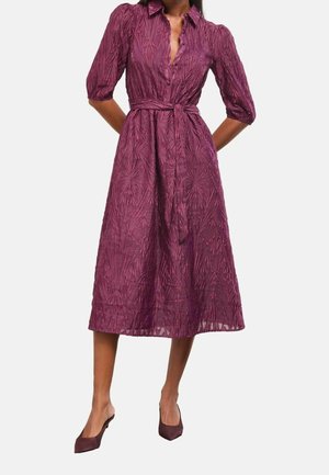 Robe midi texturée violette avec manches mi-longues, col, taille ceinturée, portée avec des talons pointus assortis, modèle debout sur un fond uni.