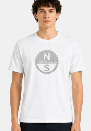 Giovane uomo che indossa una maglietta bianca con un logo circolare grigio North Sails centrato sul petto, in piedi davanti a uno sfondo semplice.