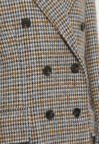 Sacou din lână, cu două rânduri de nasturi, în nuanțe de gri, negru și model pied-de-poule portocalie. Dispune de nasturi mari negri și un finisaj din țesătură texturată.