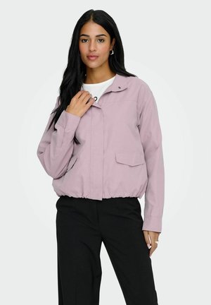 Hosszú fekete hajú nő világoslila dzsekit, fehér inget és fekete nadrágot viselve, sima háttér előtt áll.