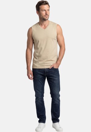 Homme portant un débardeur beige à col en V, un jean bleu foncé et des baskets blanches, debout avec une main dans la poche, regardant vers la droite.