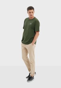 Camiseta de algodón verde oliva de manga corta, de corte relajado, con un pequeño logo en el pecho. Combinada con pantalones beige y zapatillas negras.