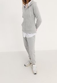 Sudadera gris con cremallera y cordones blancos, combinada con pantalones de chándal a juego. Debajo, se ve una camisa blanca con cuello. Lleva puestos unos zapatos deportivos grises.