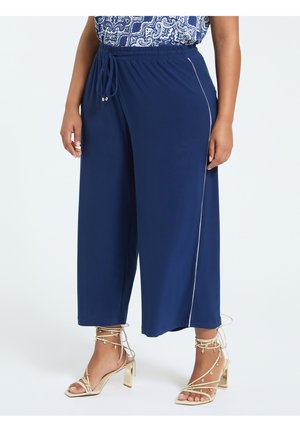 Femme portant un pantalon large bleu marine avec un liseré blanc sur le côté, associé à des sandales à talons hauts beiges à brides avec liens à la cheville.