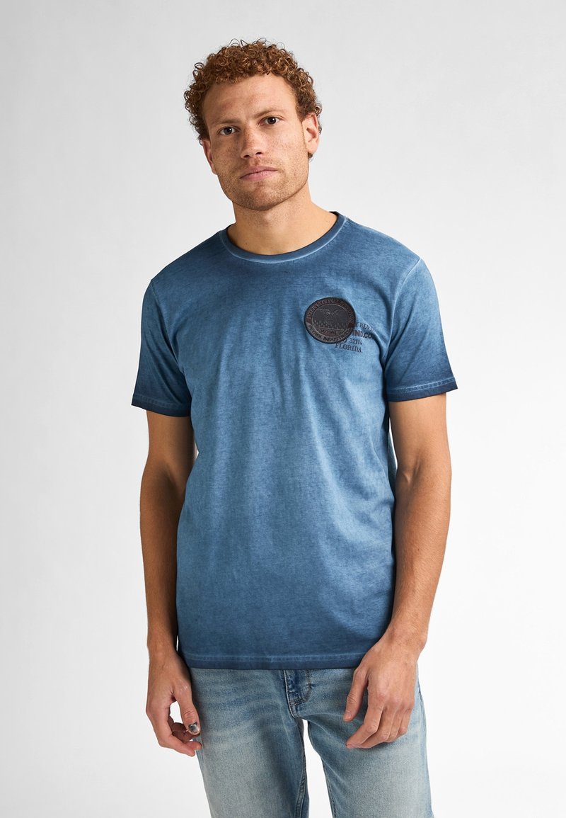 Camiseta azul de manga corta con cuello redondo y un parche circular texturizado en el lado izquierdo del pecho. La tela tiene un efecto de lavado degradado.