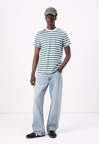 Striped t-shirt in teal and white, paired with light blue loose-fitting jeans and black sneakers. Accessorized with a gray cap.