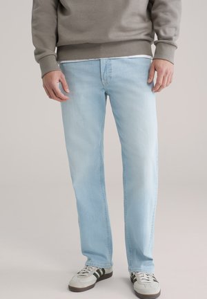 Lichtblauwe jeans met rechte pijpen, gedragen met een beige sweatshirt en witte sneakers met donkere strepen, zichtbaar van taille tot voeten.