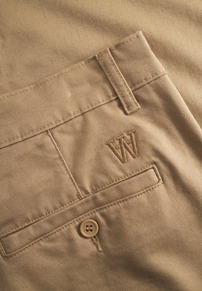 Pantaloni di tessuto beige con una tasca posteriore dotata di bottone e logo ricamato "W". La texture è liscia con dettagli di cucitura visibili.
