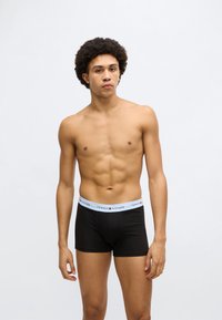 Mustad puuvillased bokserid helesinise vöökohaga, millel on "Tommy Hilfiger" logo. Modell seisab neutraalse ilmega.