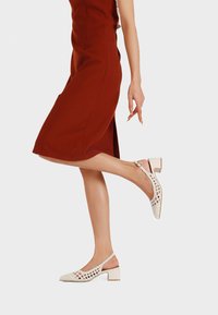 Decolleté beige con design perforato, punta quadrata e tacco a blocco. Texture liscia con una cinghia alla caviglia sicura. Indossati con un vestito rosso.