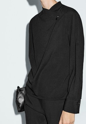 Personne portant un haut noir à manches longues avec un col asymétrique et un détail de bouton, tenant une petite pochette en cuir noir.