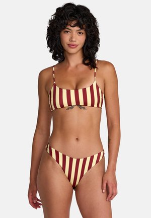 Frau mit lockigem dunklem Haar, die einen gelb und bordeauxfarben vertikal gestreiften Bikini trägt, mit einem Tattoo unter der Brust.