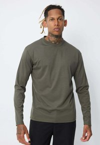 Homme avec des tatouages et des dreadlocks portant une chemise à manches longues olive et un pantalon noir, se tenant contre un fond blanc uni.