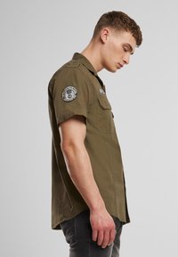 Olivengrønn kortermet skjorte med knappelukking, lapp på venstre erme og to brystlommer. Bomullsmateriale, lett teksturert.