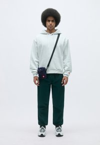 Felpa grigia in cotone con cordini, pantaloni verdi e sneakers bianche. Borsa a tracolla nera con logo rosso; il modello è in piedi contro uno sfondo neutro.