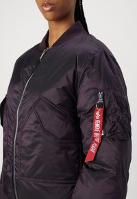 Fialová bomber bunda s předním zipem, vroubkovaným límcem a texturovaným rukávem. Obsahuje akcent klíčenky "Odstranit před odletem".