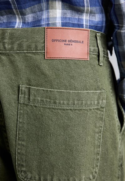 Πράσινα βαμβακερά παντελόνια με δερμάτινη ετικέτα που αναγράφει "OFFICINE GÉNÉRALE PARIS 6" στην πίσω τσέπη, με υφή και ραφές.