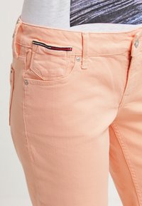 Jeans de mezclilla color durazno con una textura suave, diseño de cinco bolsillos y costuras decorativas en rojo y navy en el bolsillo delantero.