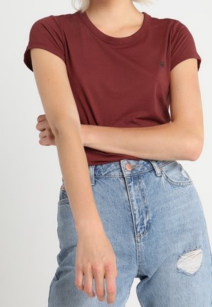 Person wearing a fitted maroon short-sleeve shirt tucked into high-waisted light blue ripped jeans, arm crossed over the torso.
