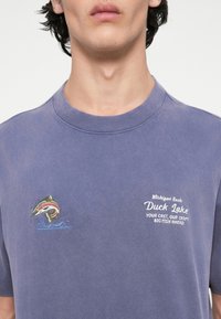 Violeta kokvilnas t-krekls ar daudzkrāsainu izšūtu zivi un baltu uzrakstu "Michigan Reels Duck Lake", papildināts ar viļņu motīviem.