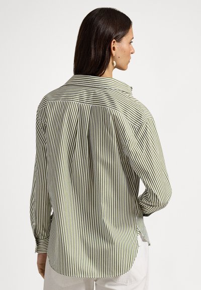 Γυναίκα με σκούρα μαλλιά που φοράει μακρυμάνικο πουκάμισο με κάθετες πράσινες και λευκές ρίγες, εμφανίζει την πλάτη της, σε συνδυασμό με λευκό παντελόνι.