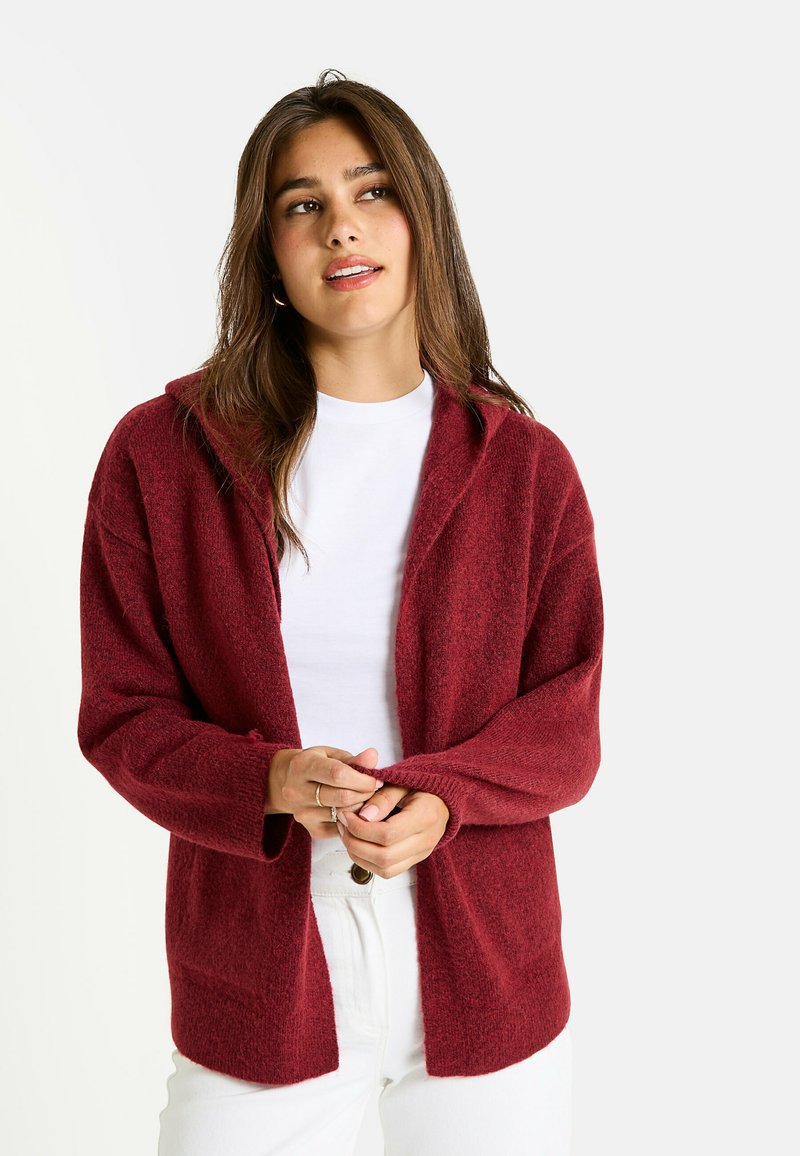 Cardigan bordeaux avec une finition douce et texturée, design à l'avant ouvert et manches longues, associé à un t-shirt blanc et un pantalon clair.