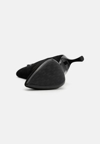 Zwarte suède stiletto-hak met een puntige neus. Glanzende zwarte zool heeft een gestructureerd grippatroon. Het ontwerp heeft een accentueerde voetboog.