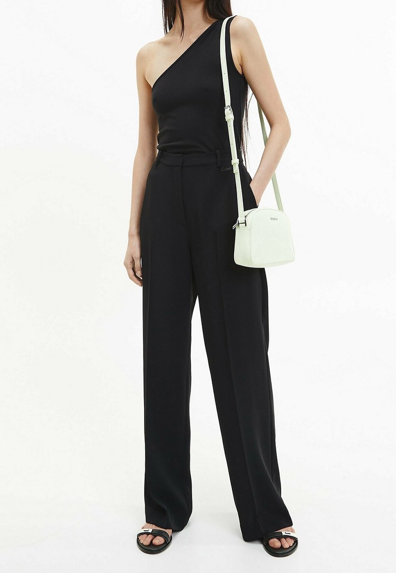 Femme portant un haut noir asymétrique, un pantalon noir ample, des sandales noires et blanches, tenant un petit sac bandoulière clair.