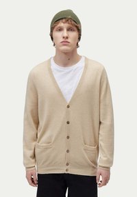 Beige kofta i mjukt material, med V-ringning, knapplist och två framfickor. Buren över en vit T-shirt.